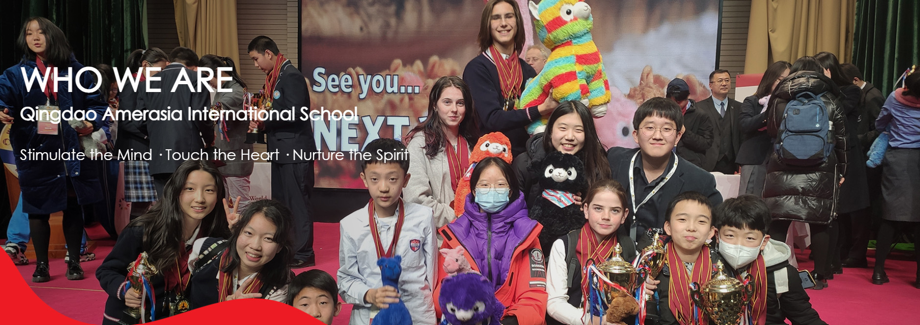 Group of students and teachers at Qingdao Amerasia International School posing proudly with trophies and colorful plush toys at a school event, under a banner reading 'Who We Are – Stimulate the Mind · Touch the Heart · Nurture the Spirit.'